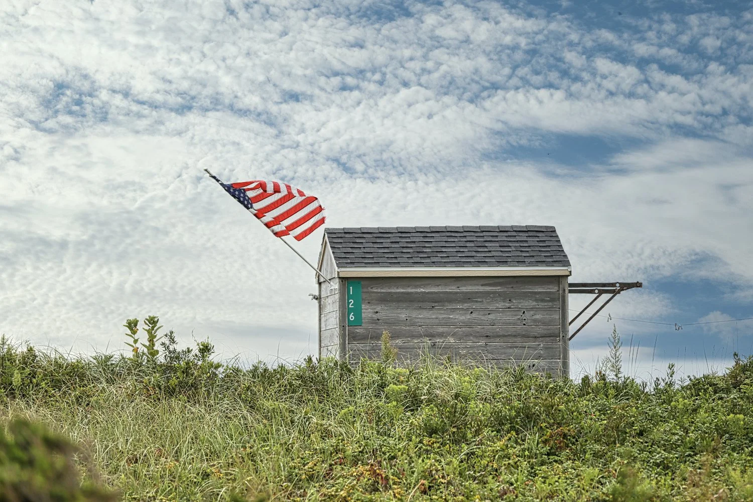 A small wooden house with a gray shingled roof and a green address plaque reading '126' on it. An American flag is mounted on the house, blowing in the wind. The house is surrounded by green grass and plants, with a partly cloudy sky overhead.