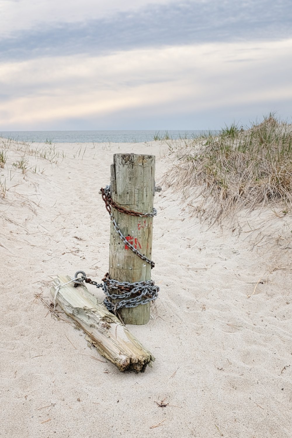 A weathered wooden post with chained metal wires on a sandy beach with dunes and grass, and the ocean and cloudy sky in the background.
