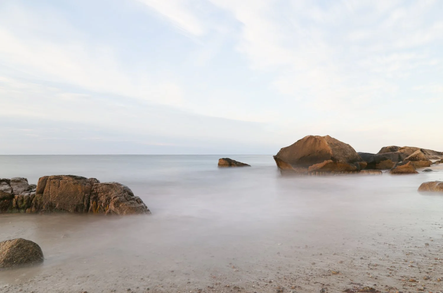 Calm ocean waters with large rocks on the sandy beach, under a partly cloudy sky.