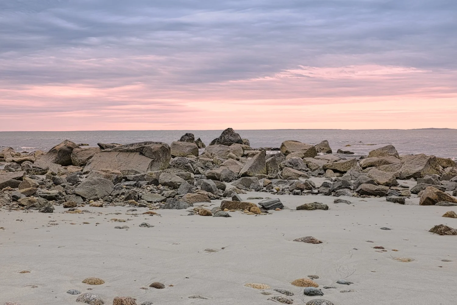 A rocky beach at sunset with a pastel pink and purple sky and calm ocean water in the background.