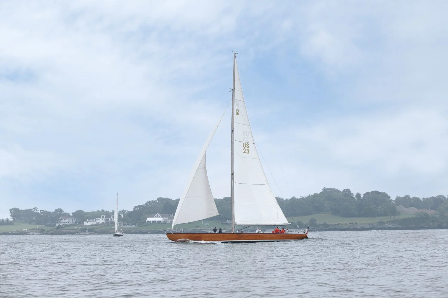 A sailboat with white sails and a wooden hull on calm water, with a shoreline of houses and trees in the background under a partly cloudy sky.