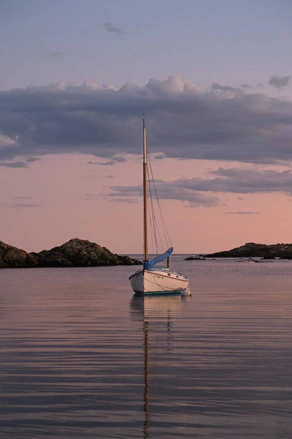 A sailboat is anchored in calm water near rocky shores during sunset or sunrise, with a pastel-colored sky and scattered clouds.