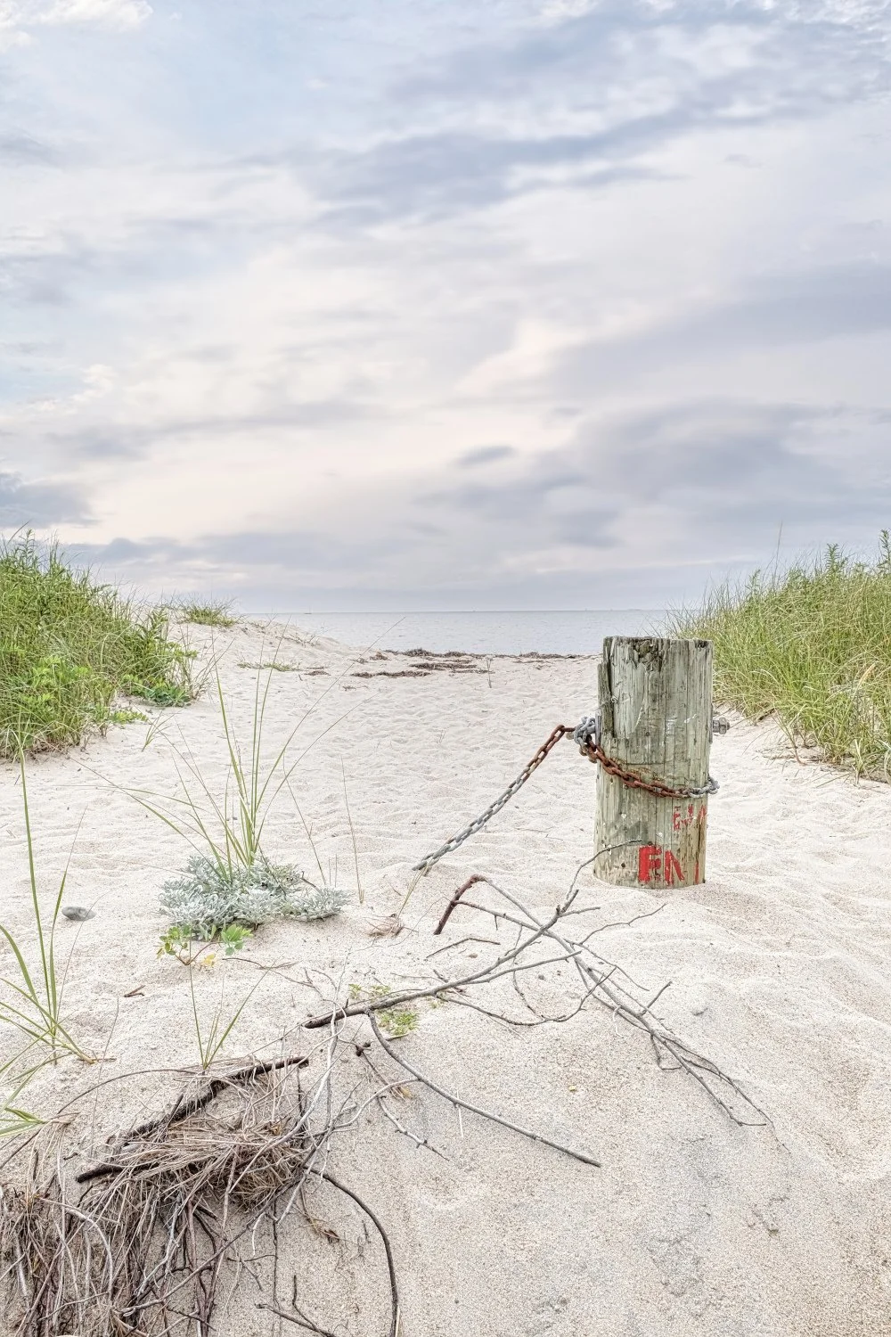 A sandy beach path bordered by grass leading to the ocean, with a weathered wooden post and rusty chain blocking the way.