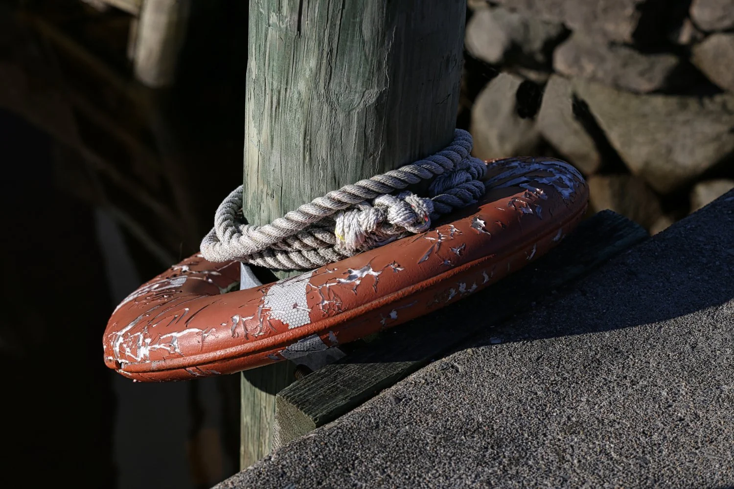 Close-up of a weathered, orange lifebuoy with peeling paint, mounted on a wooden dock post, with a gray rope looped around the post and lifebuoy, near a rocky shoreline in sunlight.