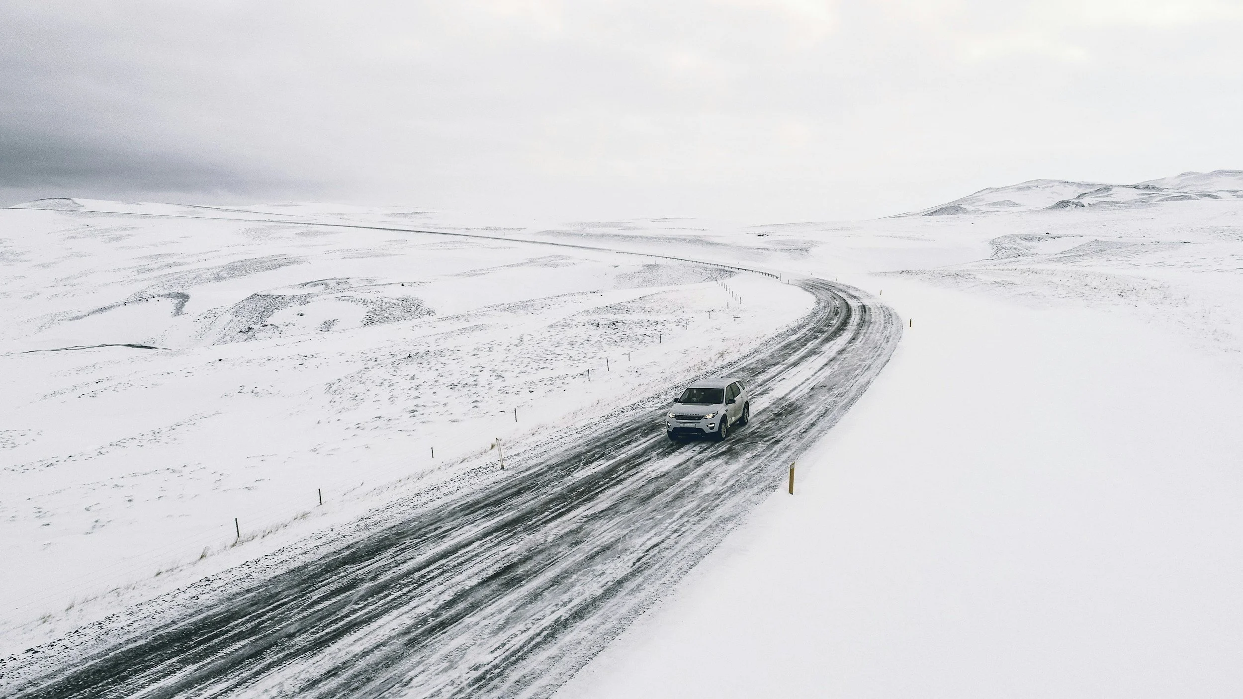 A gray SUV driving on a snow-covered mountain road with snow-blanketed hills and cloudy sky in the background.