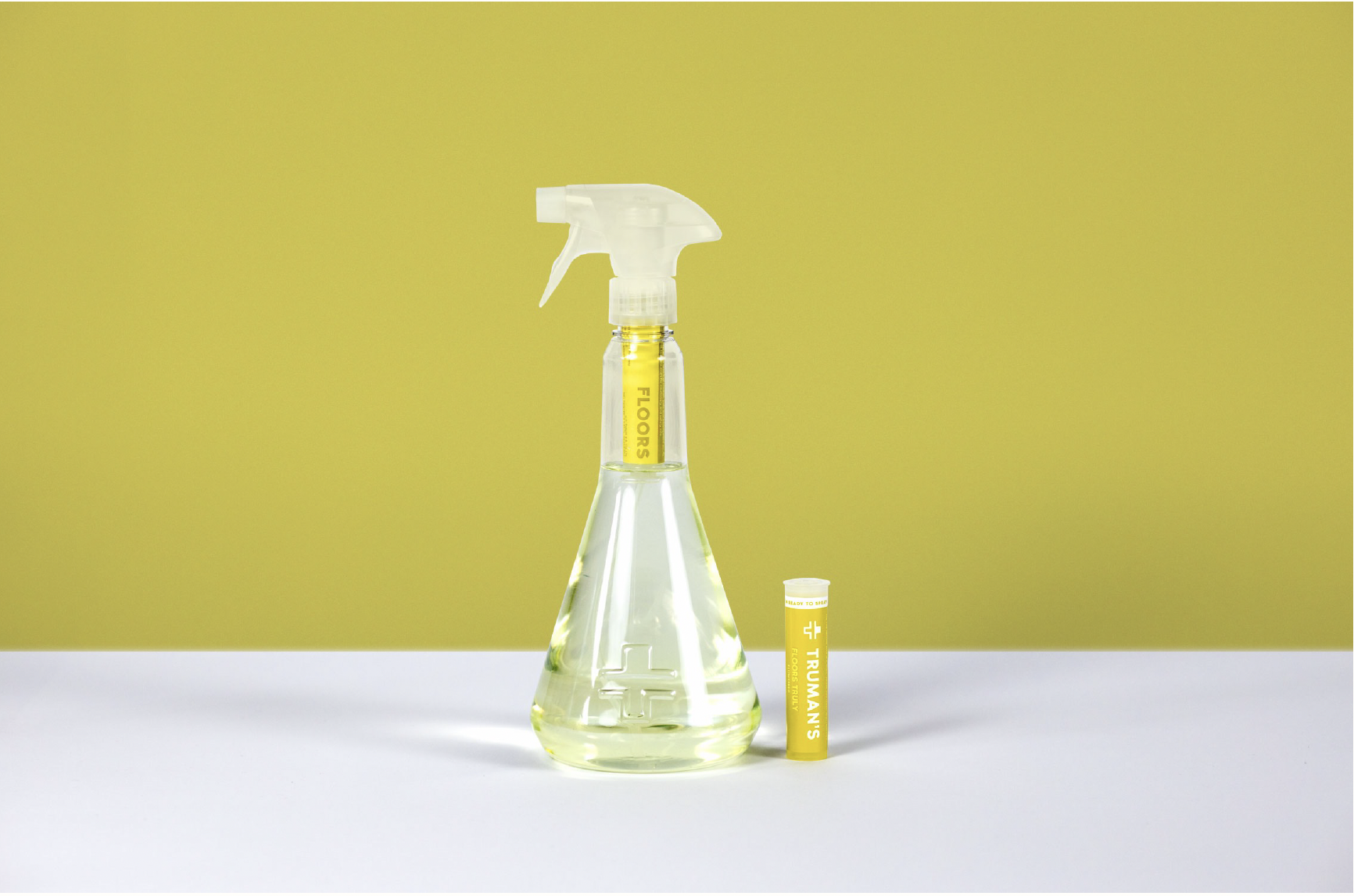 A spray bottle filled with a yellow liquid, marked 'Floors' with a spray nozzle on top. Next to the spray bottle, there is a yellow tube labeled 'Truman's Floors Truly.' The background has yellow vertical stripes, and the object is placed on a white surface.
