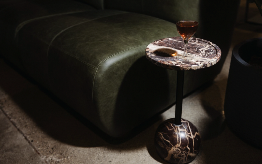 A small round table with a marble top and base, holding a glass of red wine, next to a green leather sofa in a dimly lit room.