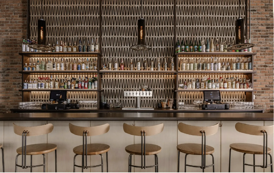 Bar with wooden shelves holding various bottles and glasses, brick wall background, four beige bar stools, black countertop, with cash registers and glassware.