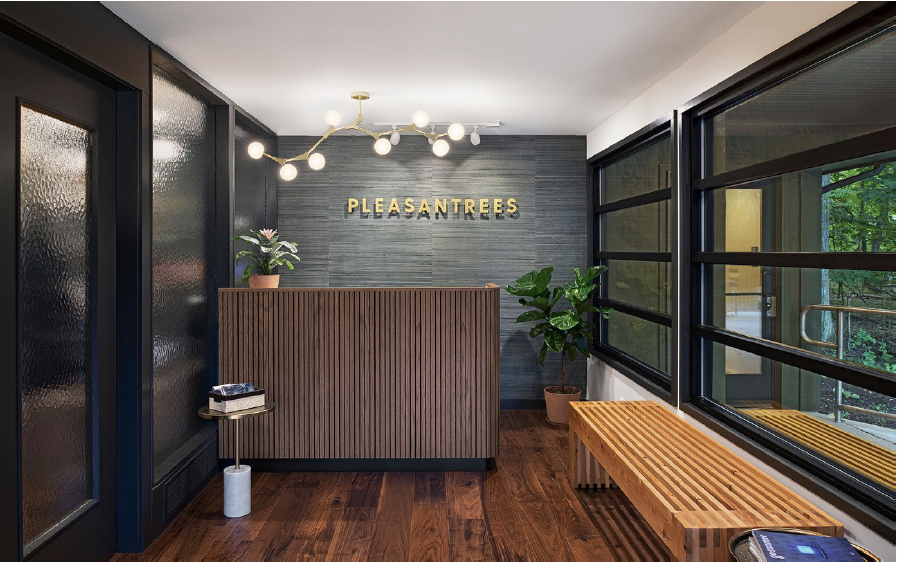 Modern office reception area with a wooden reception desk, a plant, a bench, large windows, and a sign that reads 'PLEASANTREES' on the wall.