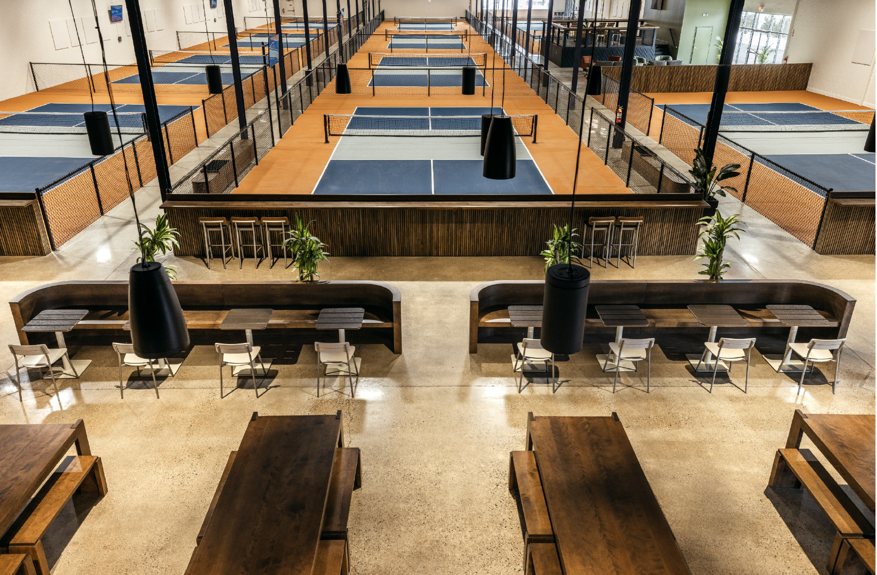 Indoor sports facility with multiple ping pong tables separated by black fences, seating areas, and large windows.