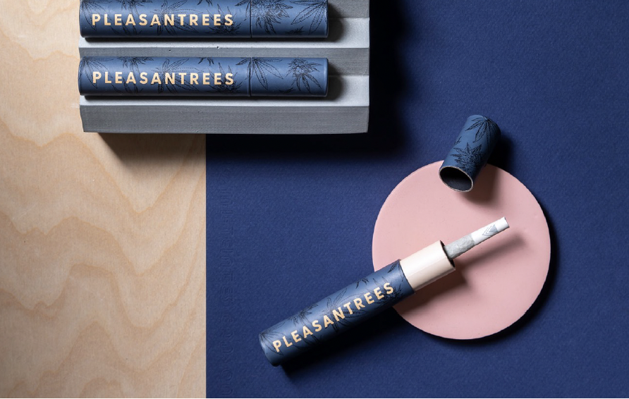 A desk scene with three blue tubes labeled 'PLEASANTREES,' a matching pen, and a pink circular object, all positioned on a blue and wood surface.
