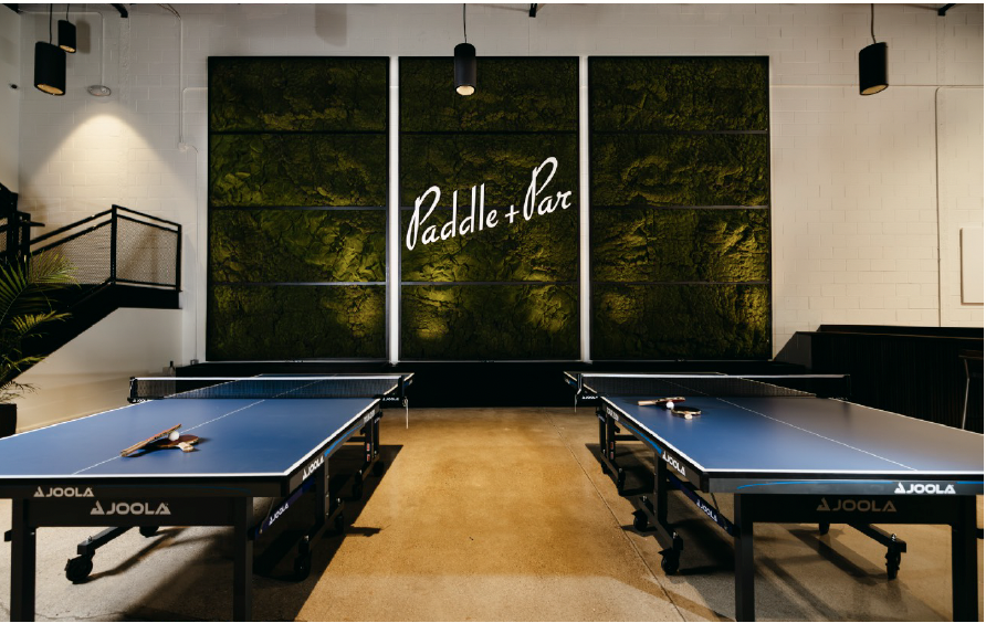 Indoor ping pong facility with two blue table tennis tables, paddles on top, and a green moss wall with a sign reading 'Paddle & Play' in the background.