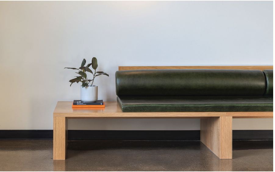 A minimalist wooden bench with a dark green cushion and backrest, placed against a light-colored wall, with a small potted plant and books on the left side.