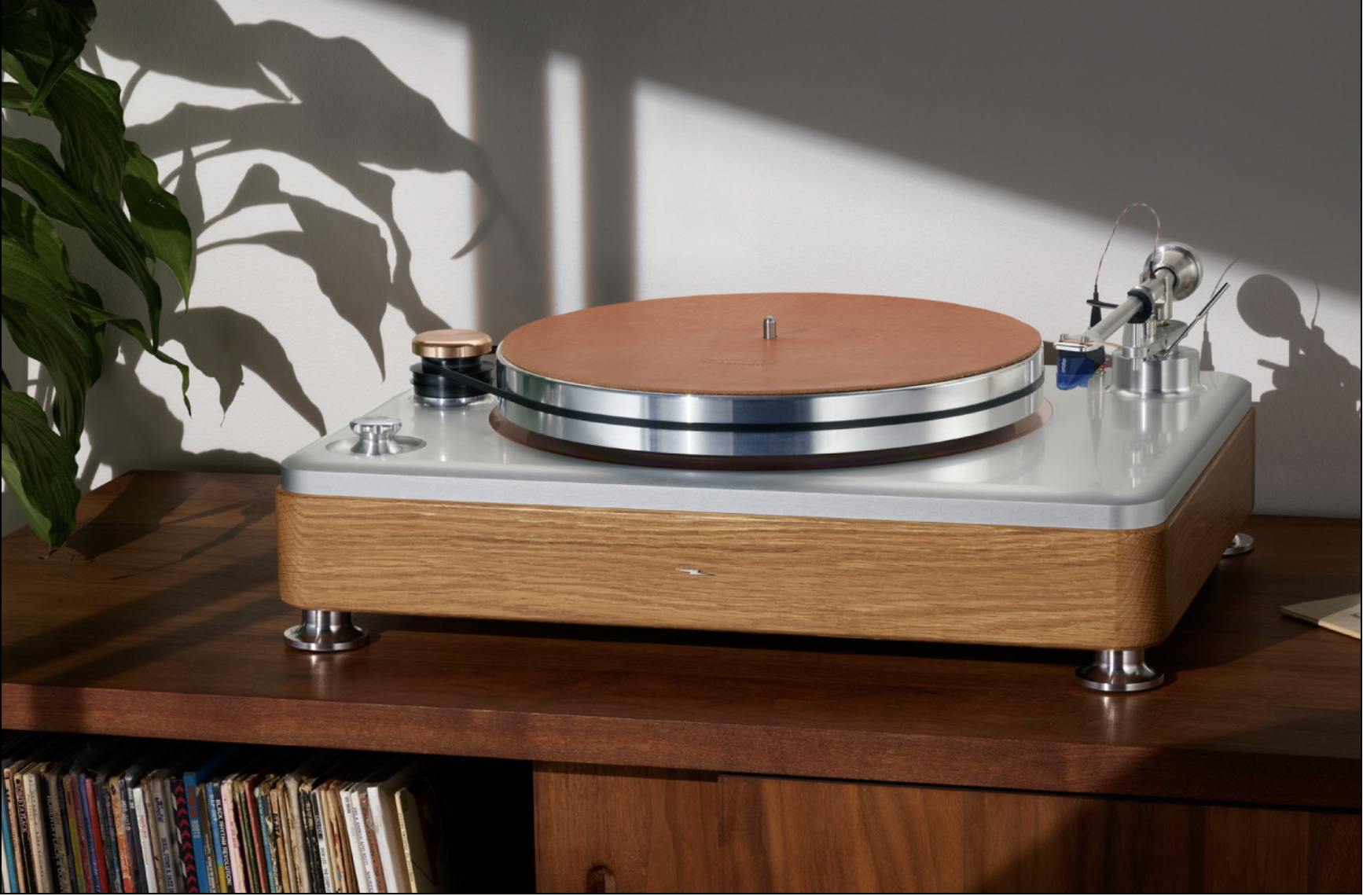 A vintage turntable with a wooden base, metal accents, and a brown platter, placed on a wooden table with a shelf of vinyl records below, and a plant casting shadows on a white wall as sunlight hits.