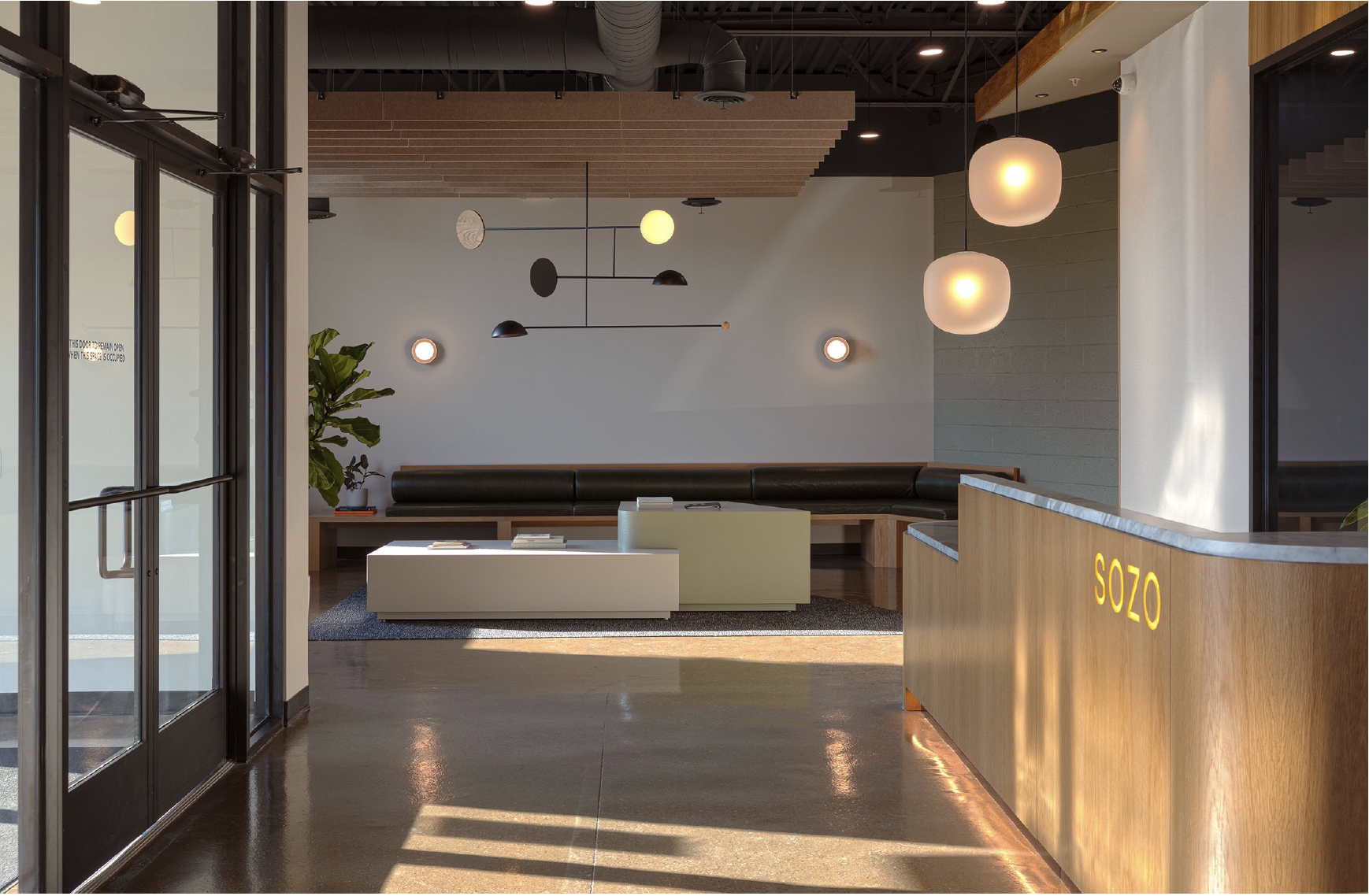 Modern hotel lobby with a reception desk labeled 'SOZO', a seating area with a black leather corner sofa, a white coffee table, and a large green plant. The space is decorated with geometric pendant lights and wall-mounted lamps, with wood accents and a dark ceiling.