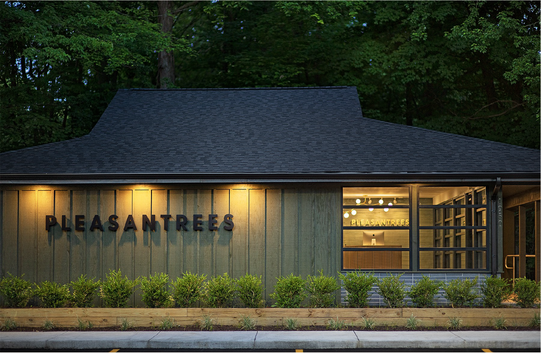 The exterior of a building with the sign 'Pleasantrees,' illuminated in warm lights, with a large window showing a reception desk and interior decor, surrounded by greenery and trees.