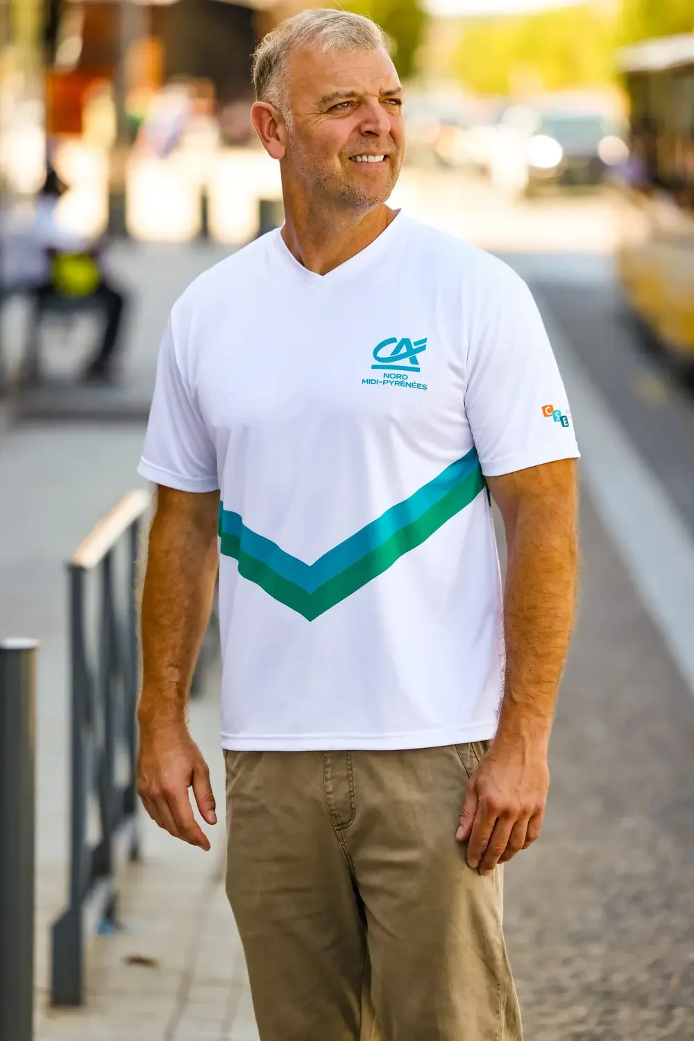 Homme souriant portant un t-shirt blanc avec logo CAF Nord Midi-Pyrénées, marche dans une rue urbaine en journée.