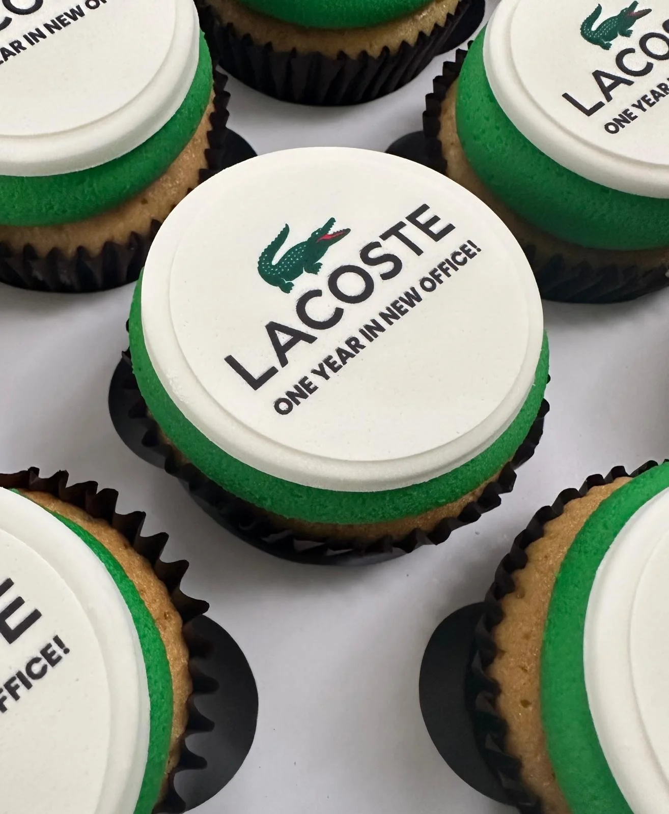 Cupcake with white fondant top, green border, and Lacoste logo featuring a crocodile and the text "LACOSTE ONE YEAR IN NEW OFFICE!".