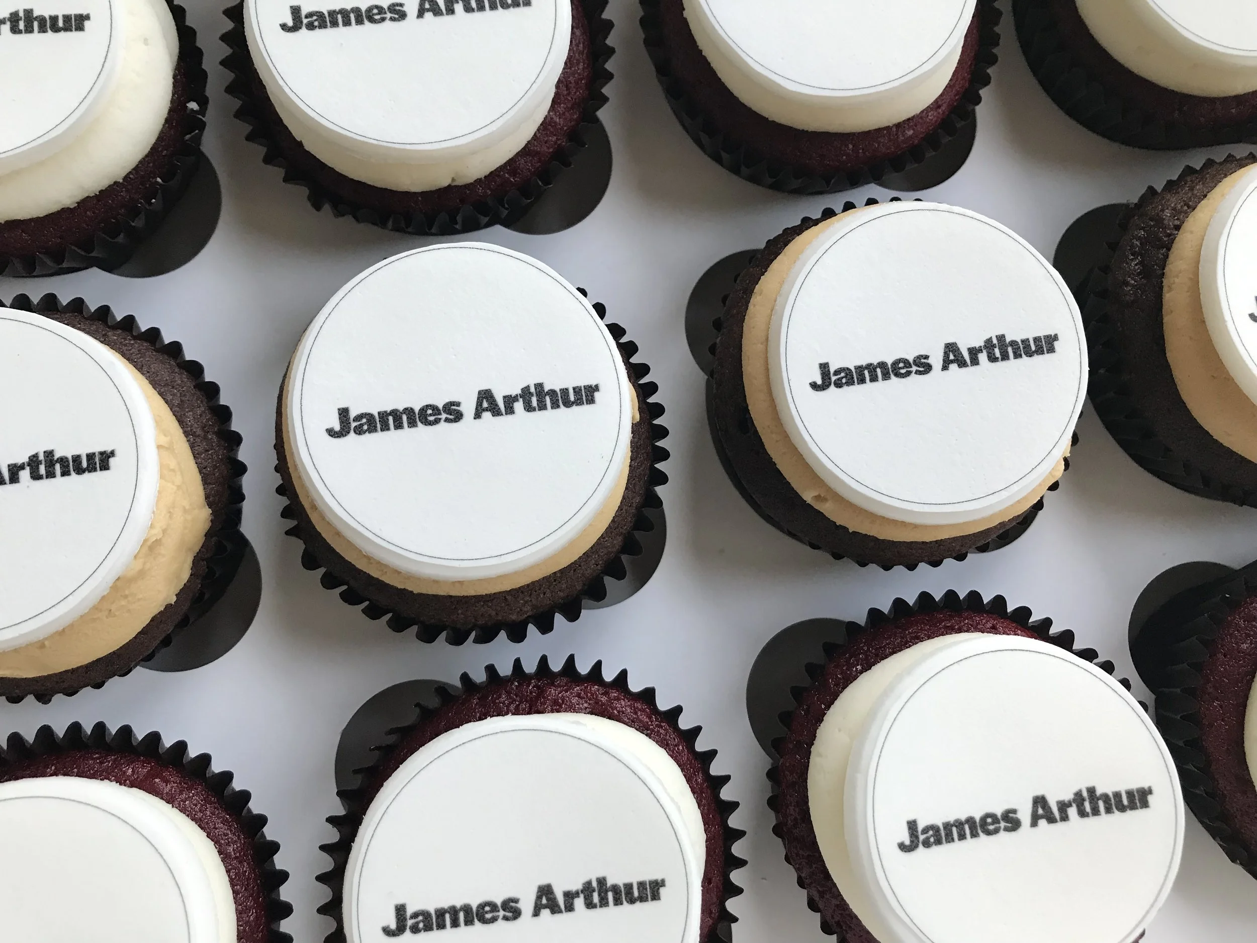 A group of cupcakes with white and dark frosting, each with a printed edible topper bearing the name "James Arthur."