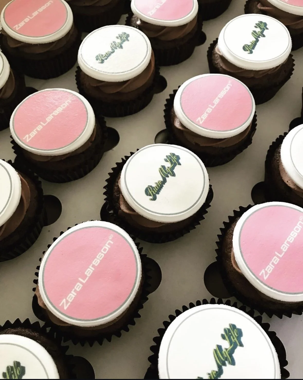 Many cupcakes in black wrappers with pink and white edible toppers that read 'ZeroLarger.com' and 'Pure Life'.