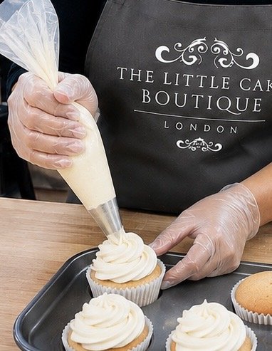 Person wearing a black apron with 'The Little Cake Boutique London' printed on it, piping white frosting onto cupcakes using a piping bag.