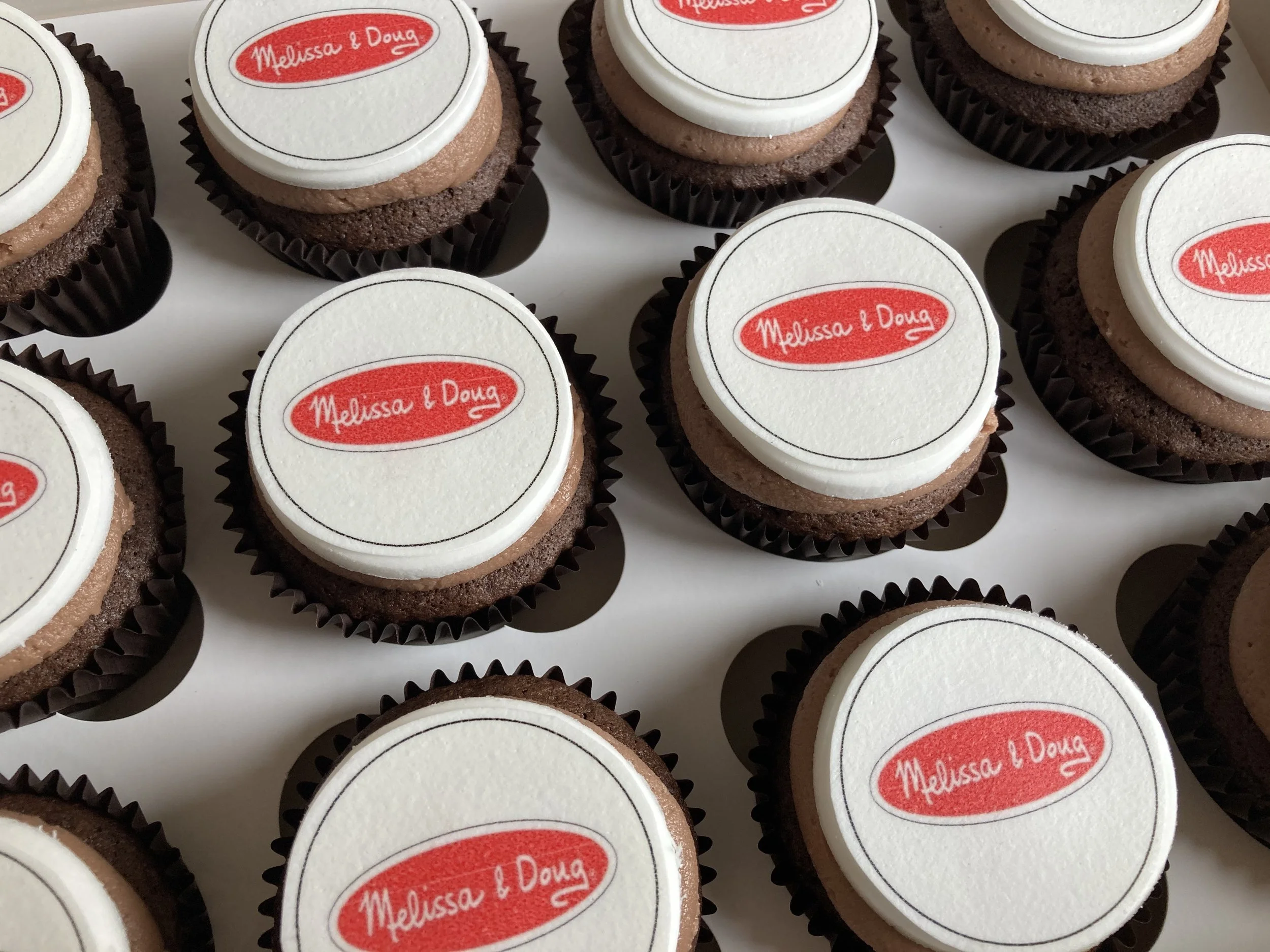 Multiple chocolate cupcakes with white fondant toppers that have red oval logos reading 'Melissa & Doug' printed on them.