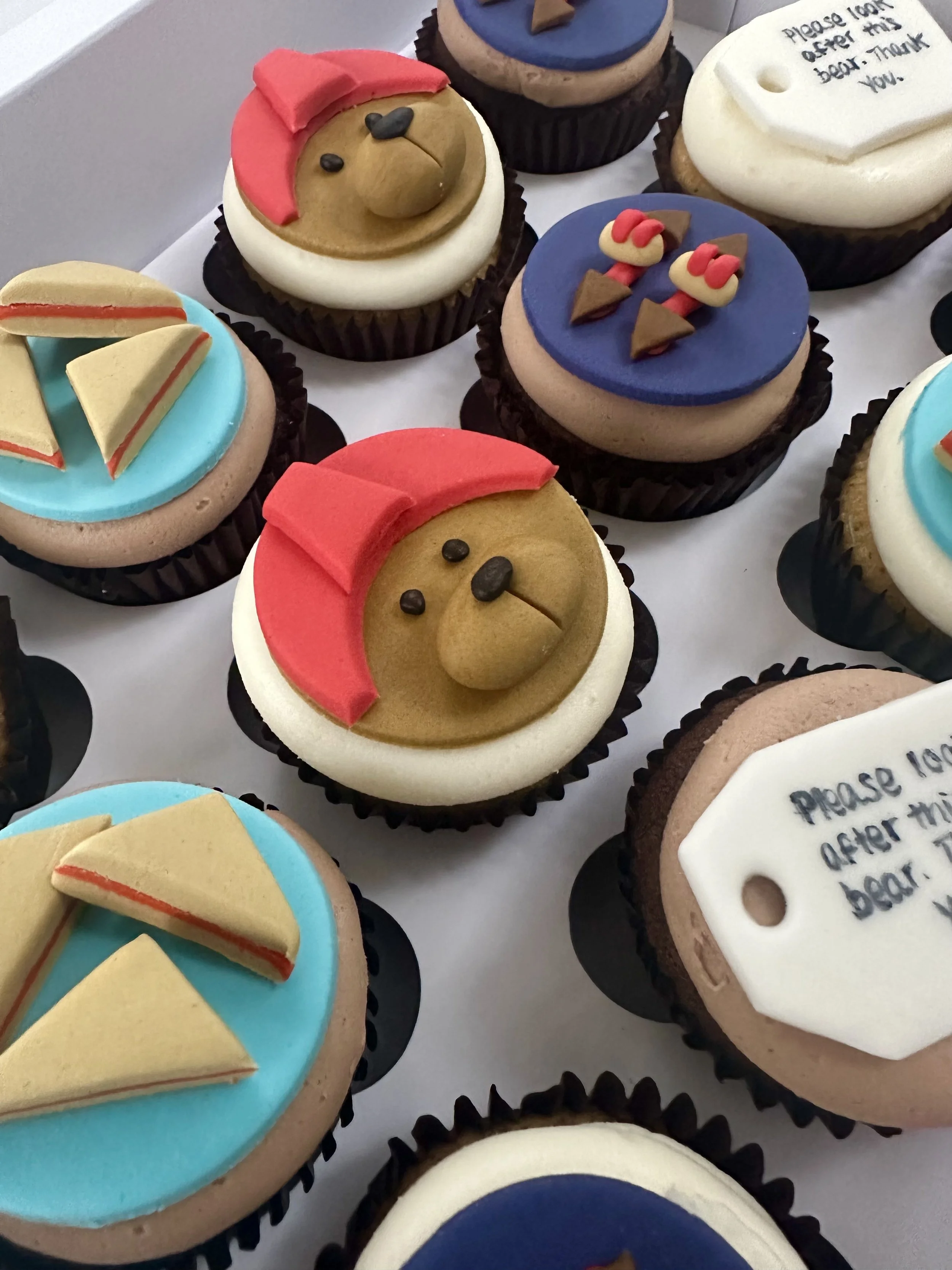 Decorative cupcakes with various themes, including a teddy bear with a red hat, a pair of summer sandals, and a cupcake with a tag that says, "Please look after this bear. Thank you."
