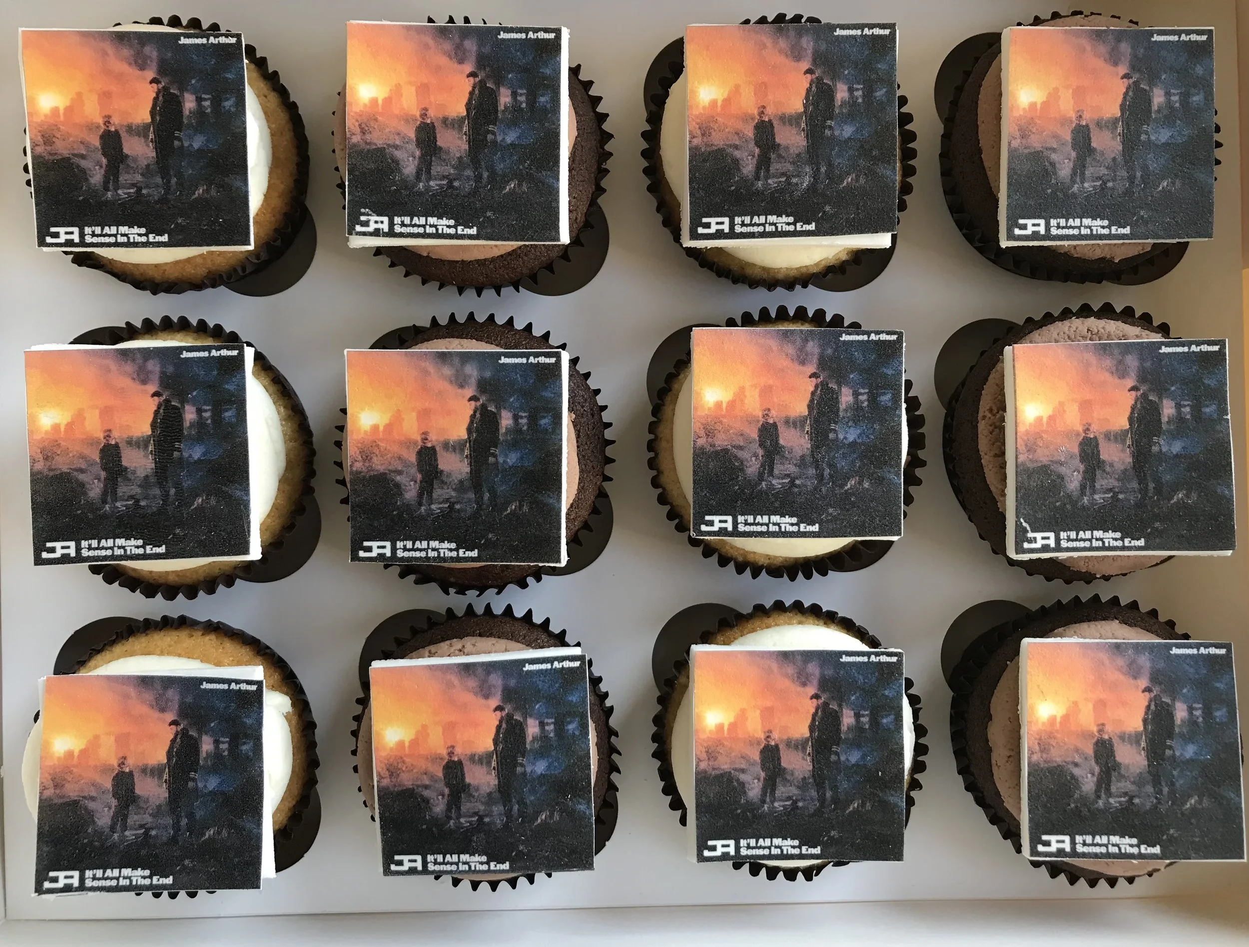 A display of cupcakes with edible images on top depicting a sunset scene with two silhouetted figures and industrial structures. The cupcakes are arranged in rows.