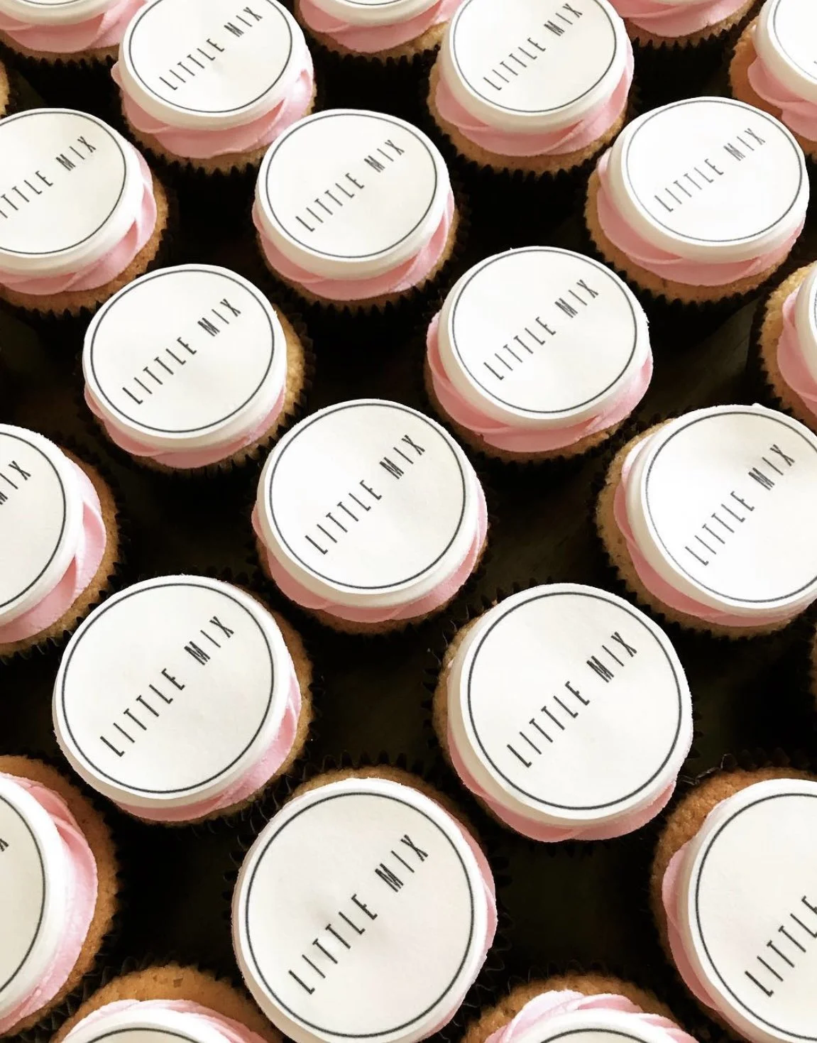 A tray of cupcakes with pink frosting, topped with white circular edible toppers that have 'LITTLE MILK' written in black.