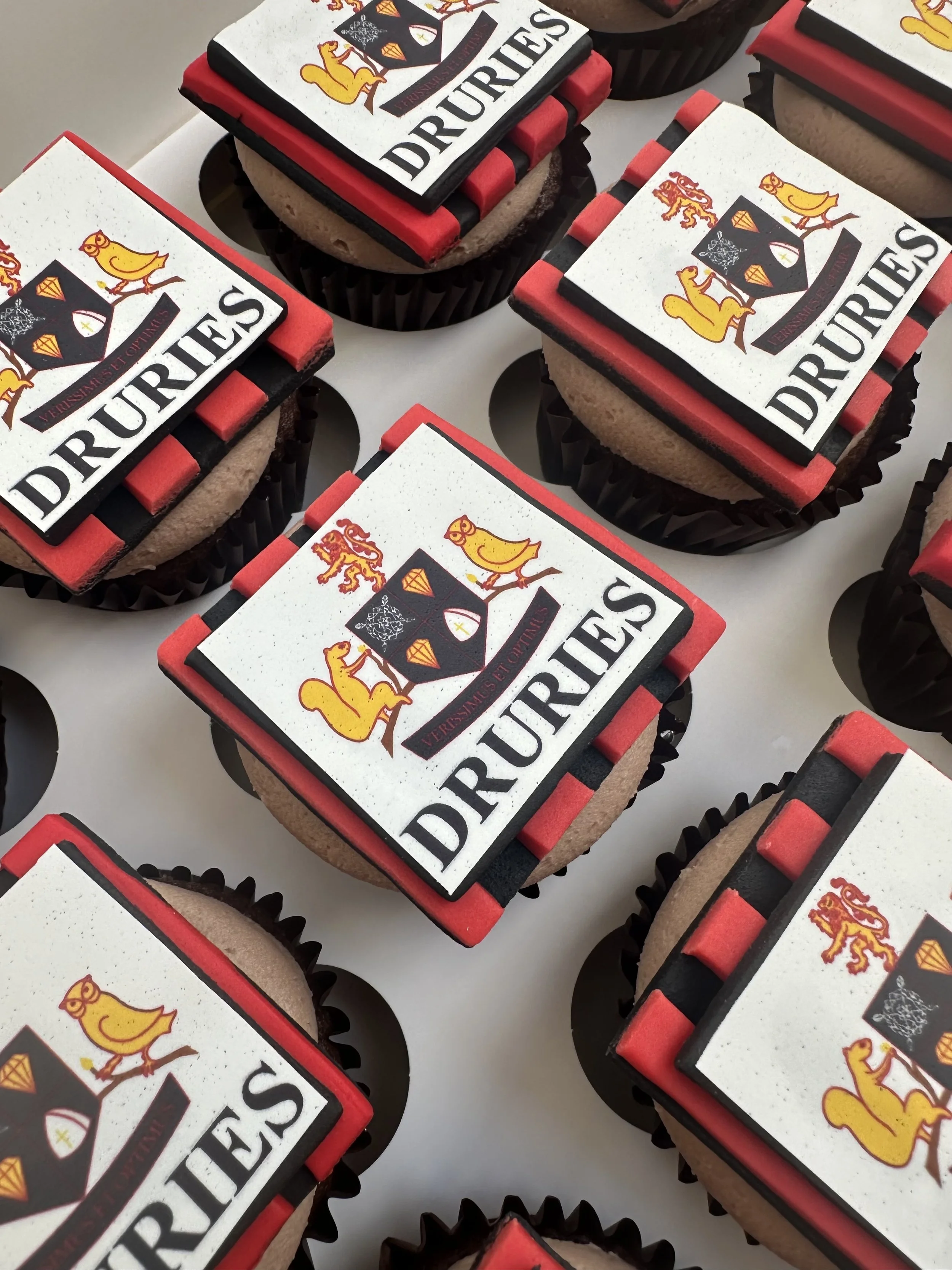 Close-up of multiple decorated cupcakes with a Druries logo on edible toppers featuring a yellow dog and a black shield.