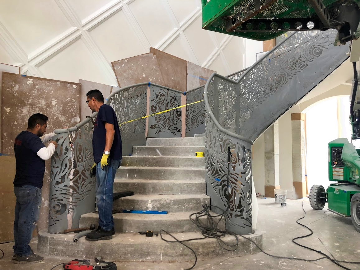 Two workers installing decorative metal railings on a staircase in an interior space, with construction equipment and unfinished walls around them.