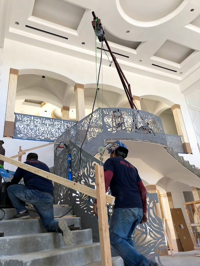Construction workers installing decorative metal panels on a staircase in an indoor building with a vaulted ceiling.