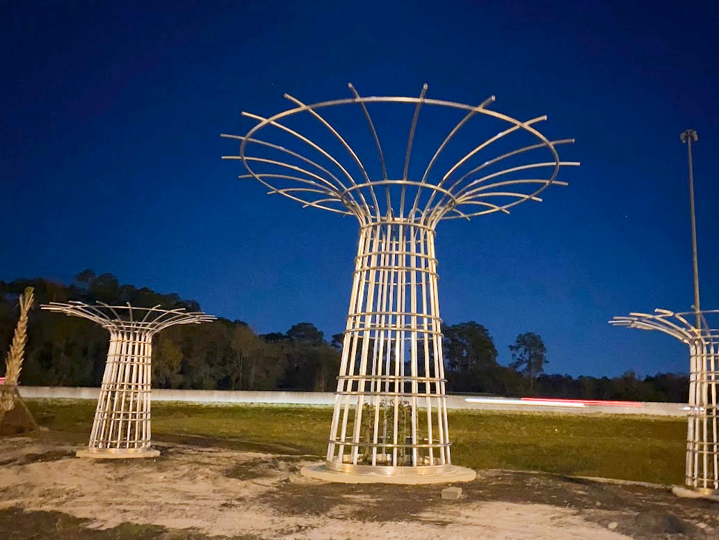 Metal tree sculptures at dusk with dark blue sky and faint tree line in the background.