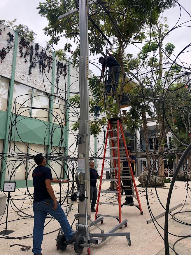 People assembling or repairing a large, circular art installation with intertwined black wires outdoors, with a Tree and building with Gucci signage in the background.