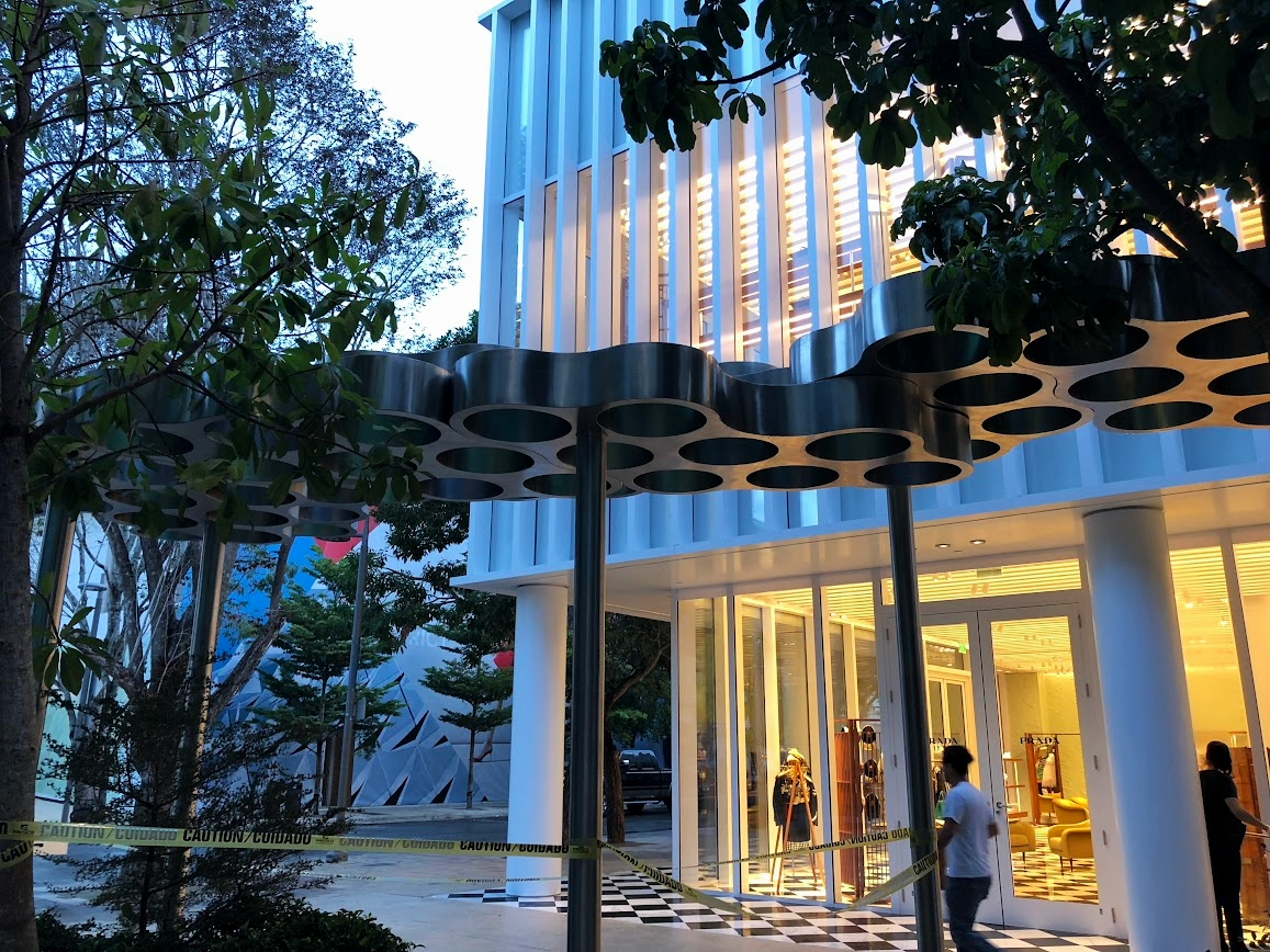 Exterior view of a modern building with large glass windows, illuminated from inside, with trees and a tree-shaped sculpture outside, and caution tape on the ground indicating construction or maintenance.