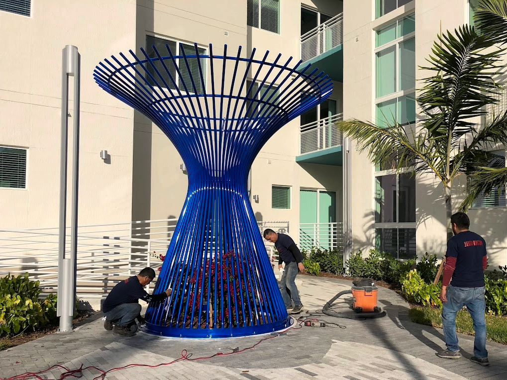 Three workers installing a large, blue, modern outdoor sculpture resembling an inverted cone with vertical bars.