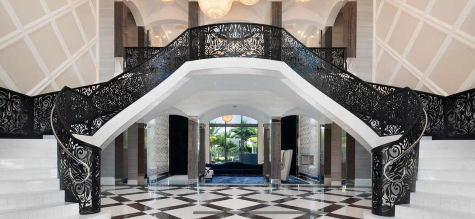 Interior view of a luxurious building with a grand staircase featuring ornate black railings, marble floors with a black and white checker pattern, and large windows or glass doors revealing palm trees outside.