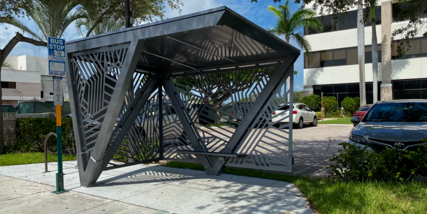 Modern bus stop shelter with geometric metal design on sidewalk in an urban area, with parking lot, cars, trees, and a building in the background.