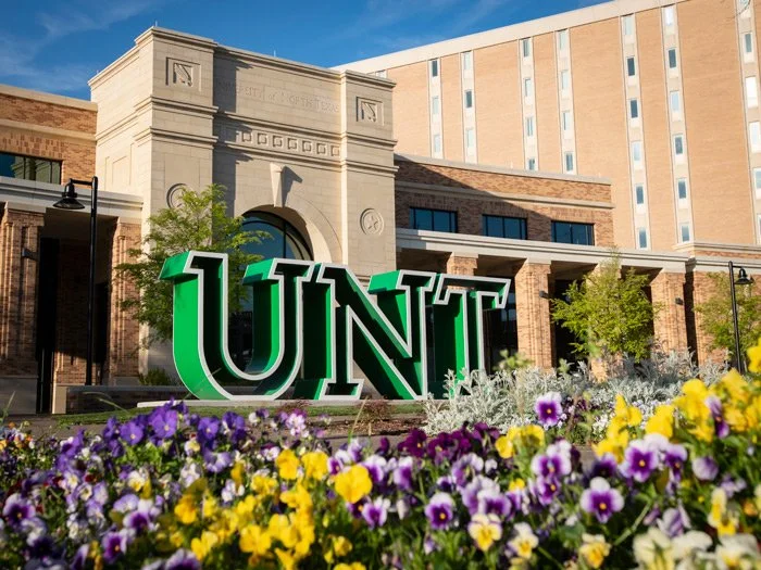 University of North Texas campus entrance with large green UNT sign, surrounded by colorful flower beds and a historic-looking building in the background.