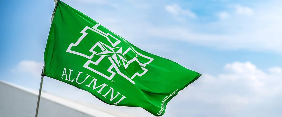 Green flag with the University of Alabama at Birmingham (UAB) Alumni logo blowing in the wind against a blue sky.