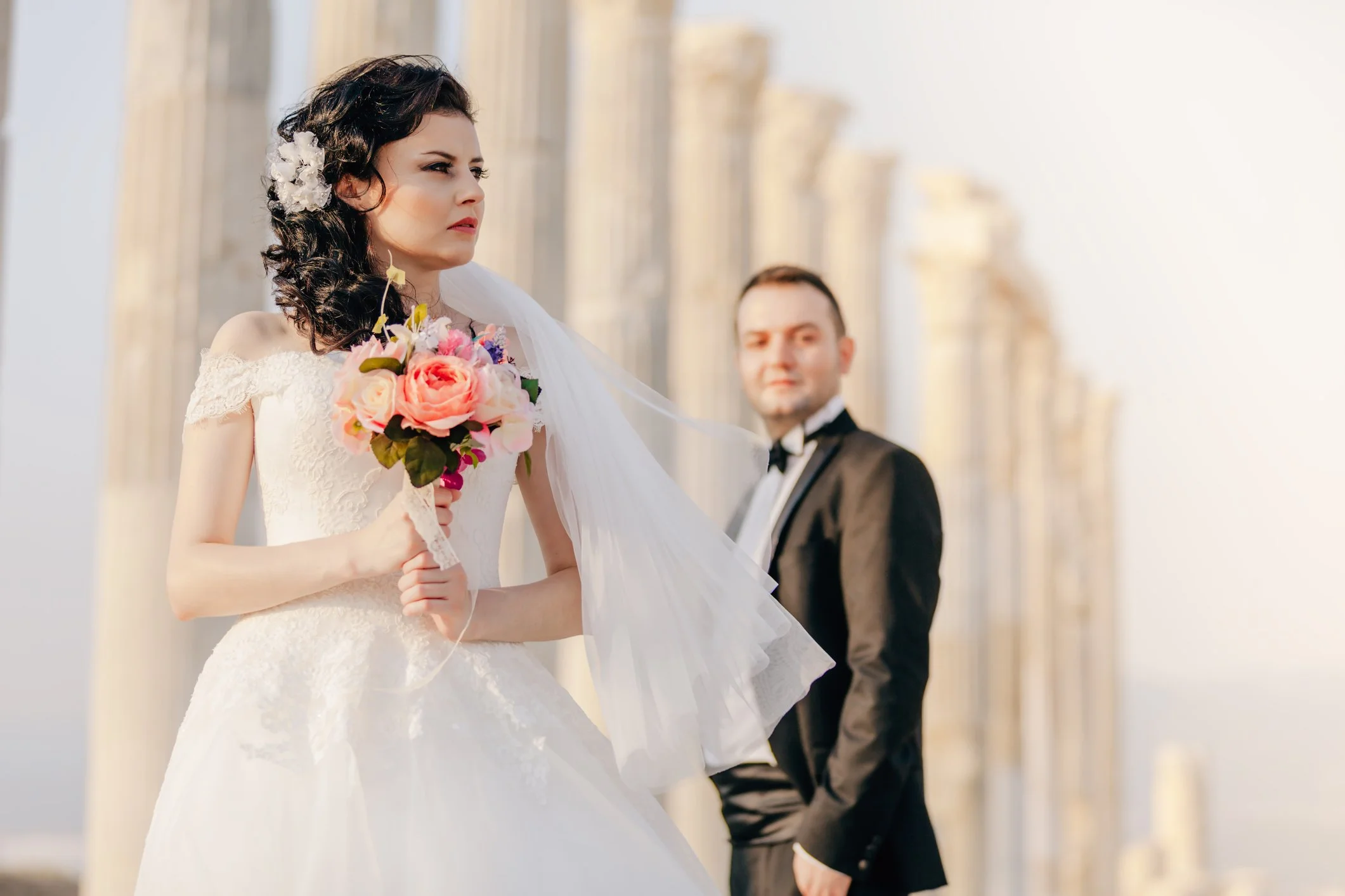 A bride in a white wedding dress holding a bouquet of pink and purple flowers, with dark curly hair adorned with a white floral hairpiece, standing outdoors with a groom in a black tuxedo blurred in the background.