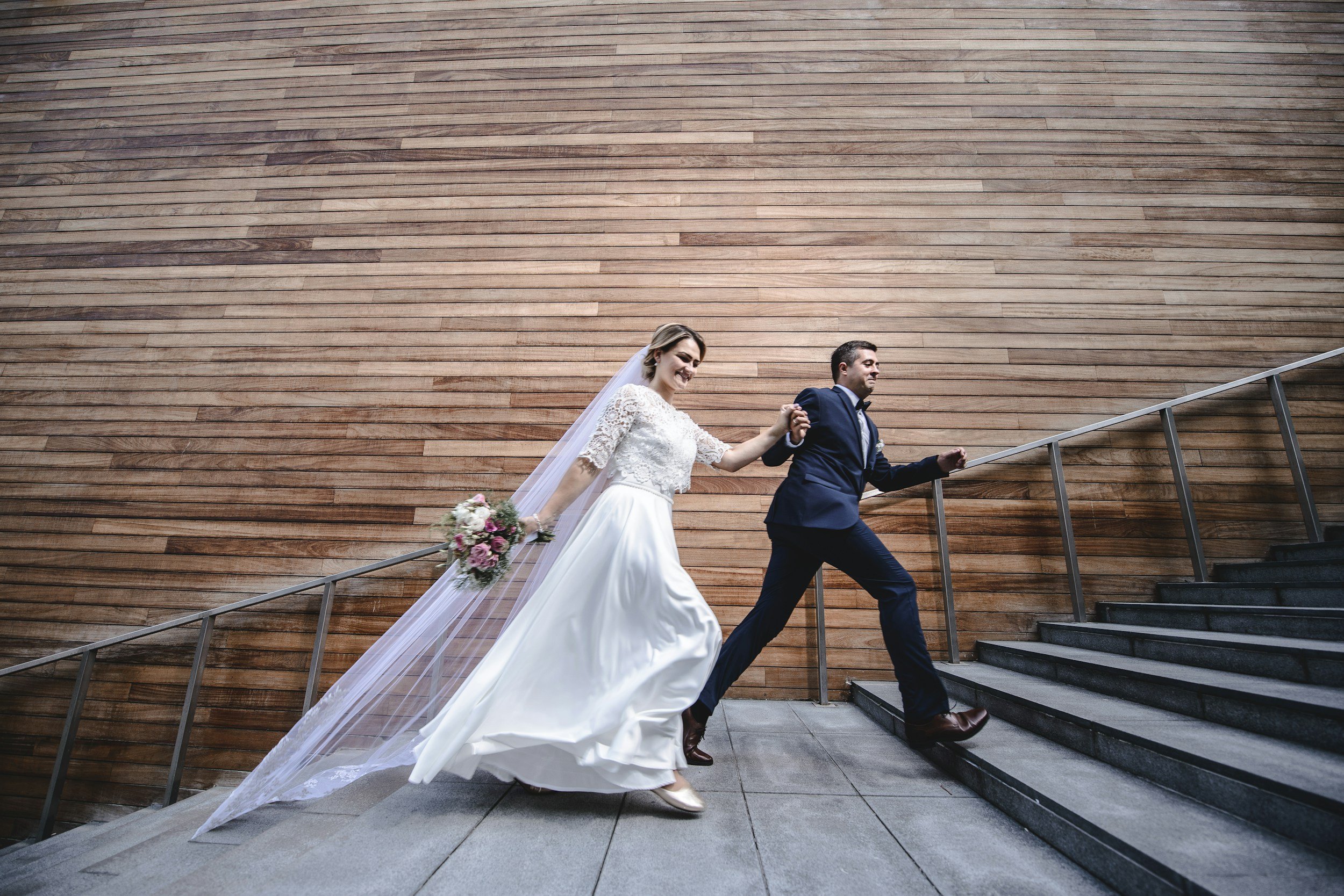 A bride and groom walking up stairs holding hands, with the bride holding a bouquet, against a wooden wall background.