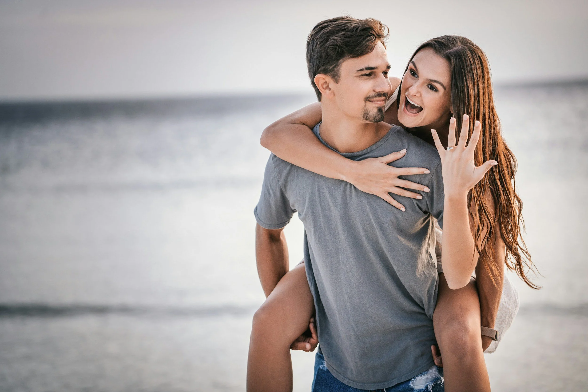 A happy couple on the beach. The woman is on the man's back, showing her engagement ring, both smiling and enjoying each other's company.