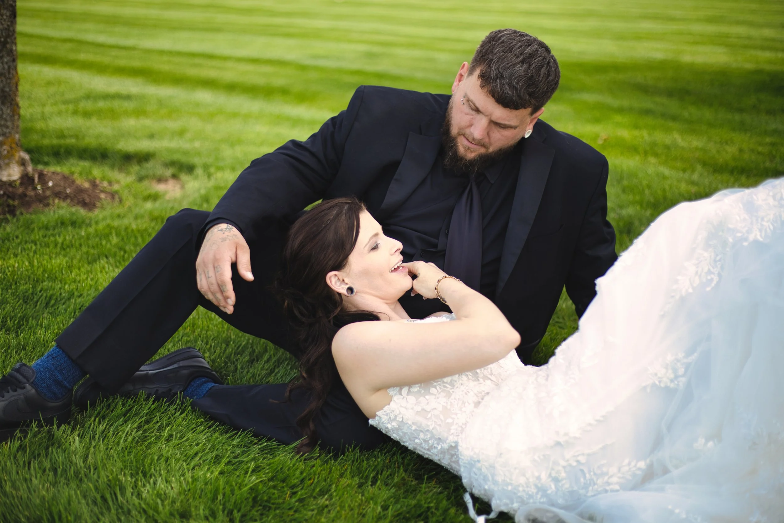 A man in black suit and a woman in a white wedding dress lying on green grass, outdoors, during a wedding photoshoot.