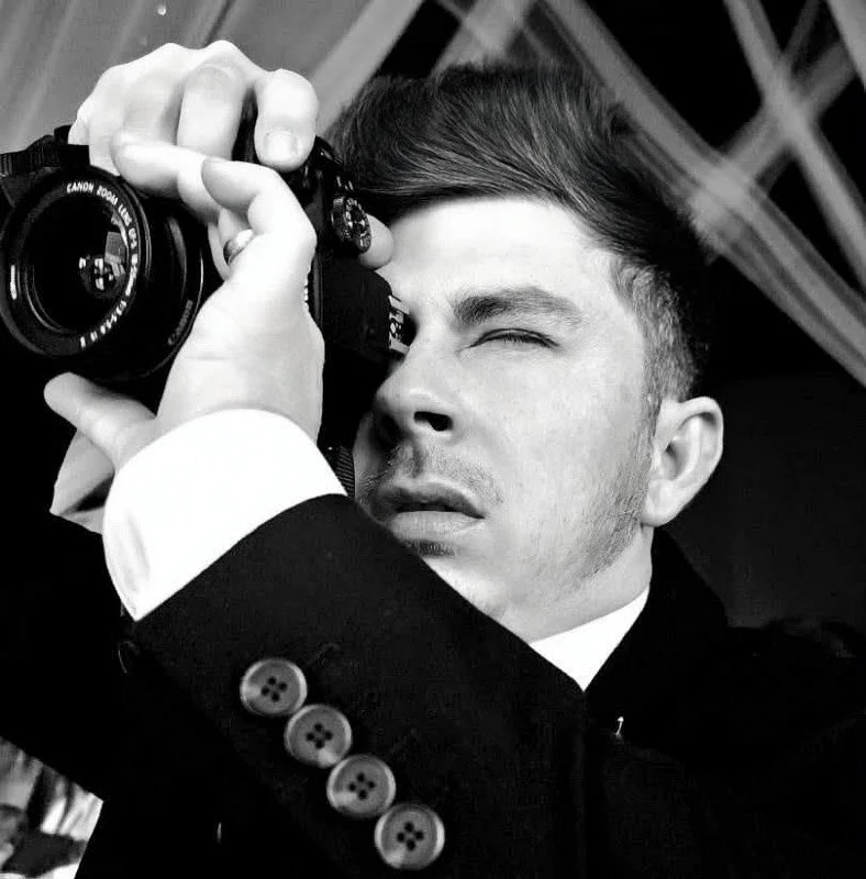 A man in formal attire holding a camera close to his face, with one eye closed as he looks through the viewfinder.