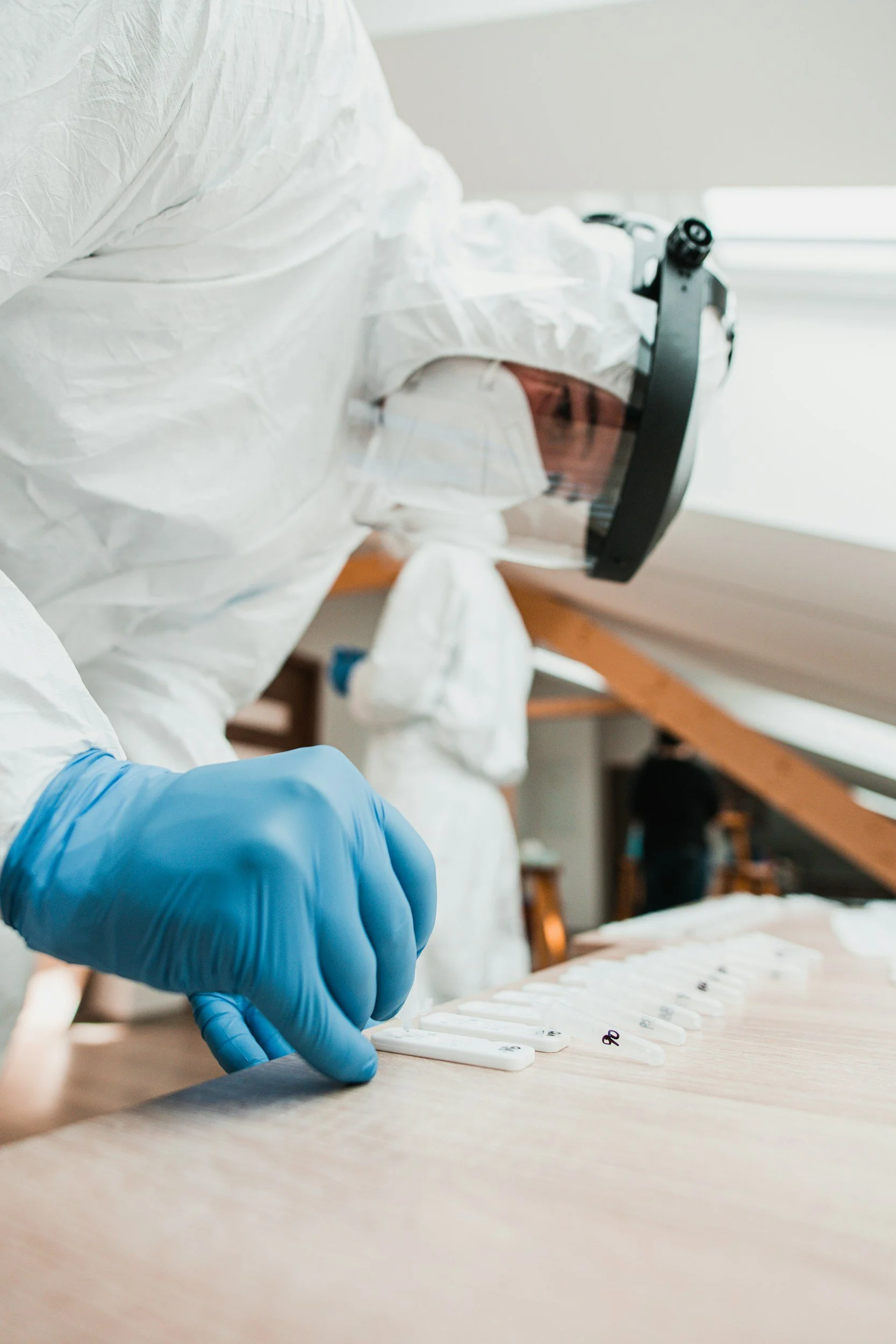 A person wearing a white protective suit, goggles, and a blue glove handling testing samples for COVID-19 or similar tests on a wooden surface with test tubes and test strips. In the background, there is a bright window and another person is faintly visible.