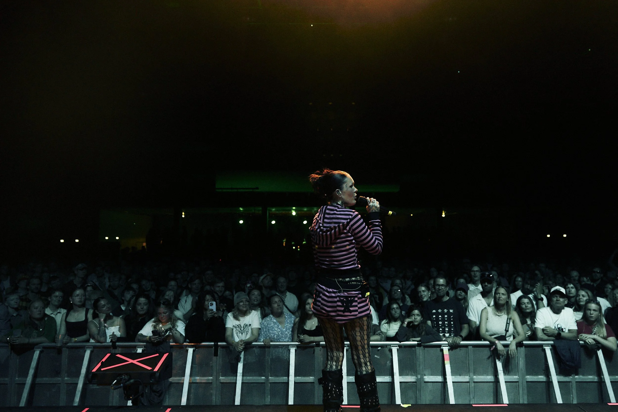 A female singer with red hair tied back, wearing a striped pink and black hoodie, fishnet stockings, and black boots, performing on stage with an audience watching in a dark, indoor concert venue.
