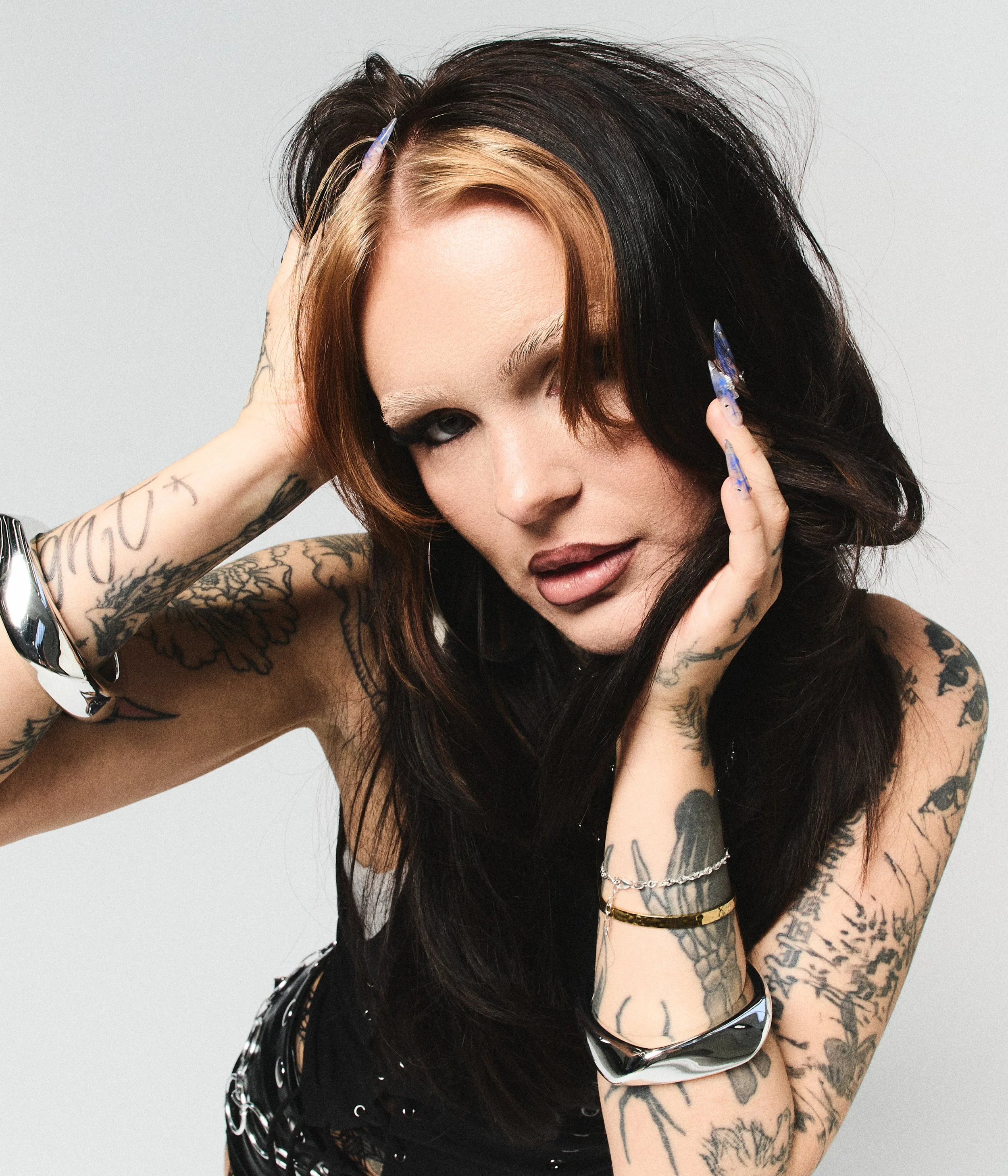 A woman with dark brown and blonde hair, multiple tattoos on her arms, and wearing a black top and silver jewelry, posing with her eyes closed and hands on her head against a plain light grey background.