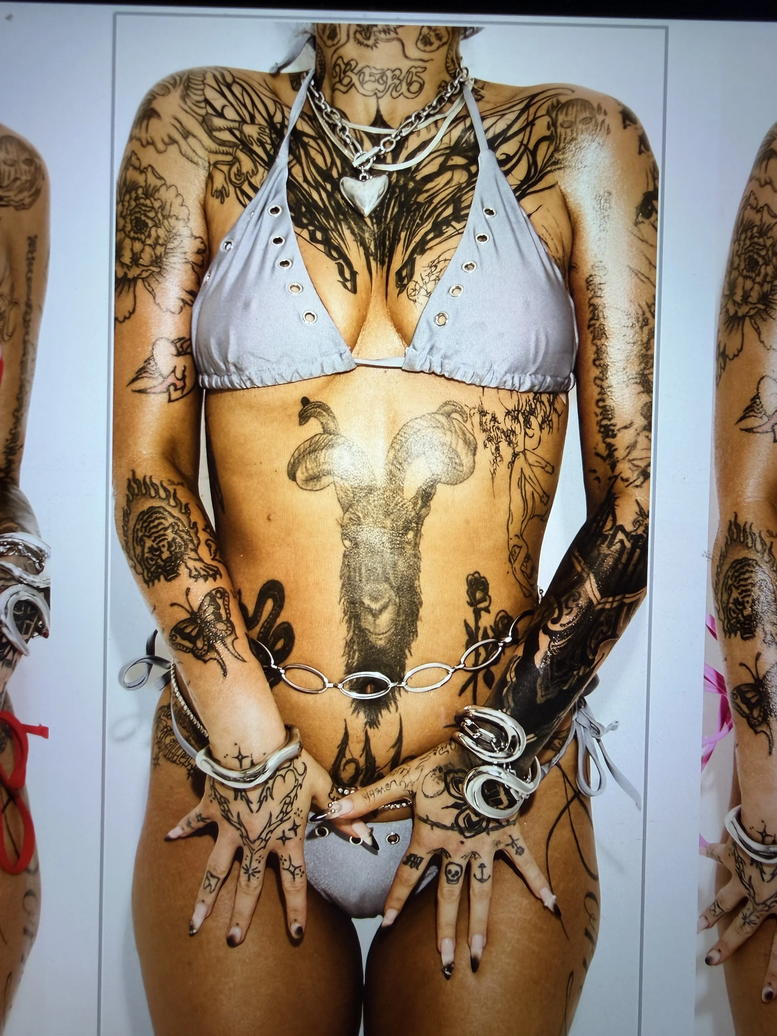 A person with numerous tattoos wearing a bikini top and jewelry, covering their private area with their hands.
