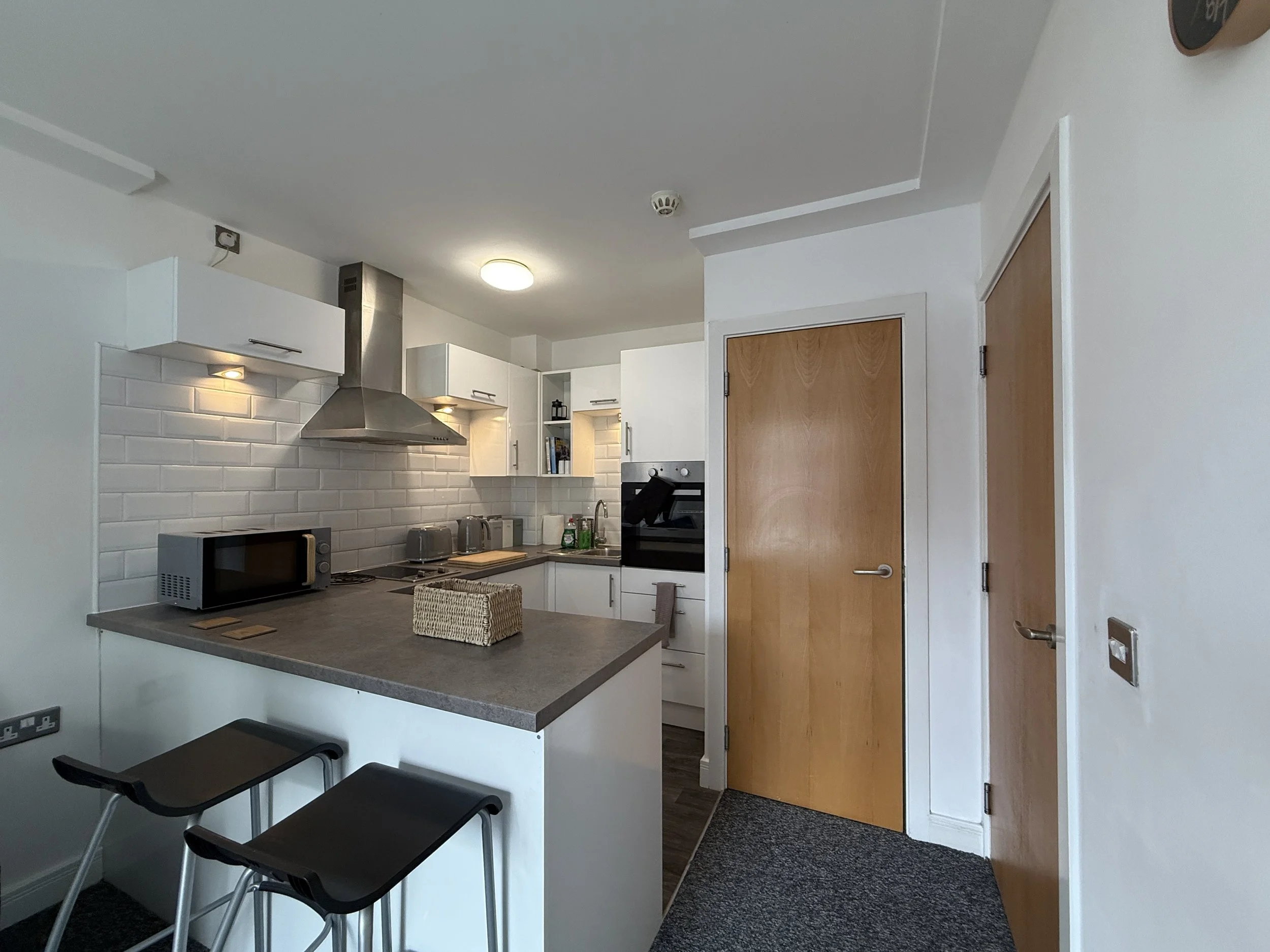 Modern kitchen with white cabinetry, grey countertops, stainless steel range hood, white subway tile backsplash, microwave oven, and a small eating area with black stools.